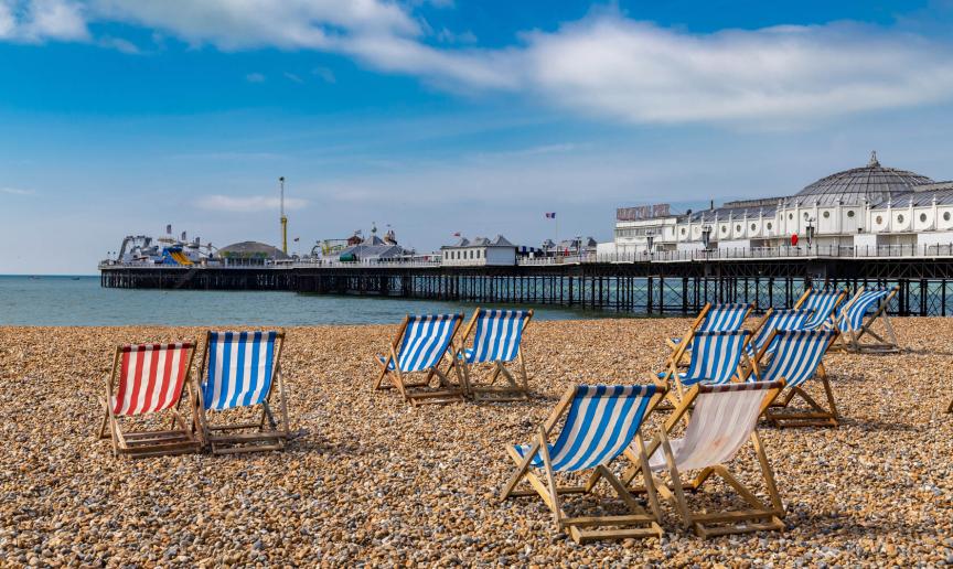Striped wooden deck chairs placed in pairs on a pebbled beach facing out towards Brighton pier and the sea.