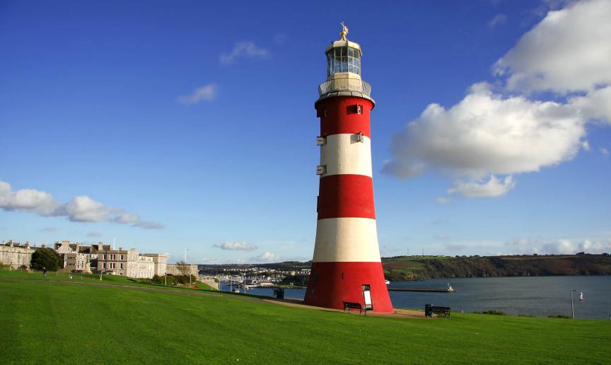 A bold red and white striped lighthouse placed on a grassy cliff, overlooking the Plymouth sound and city.