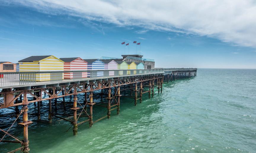 Multiple vibrant pastel and white striped beach huts lining either side of the Hastings pier, contrasting with the teal sea.