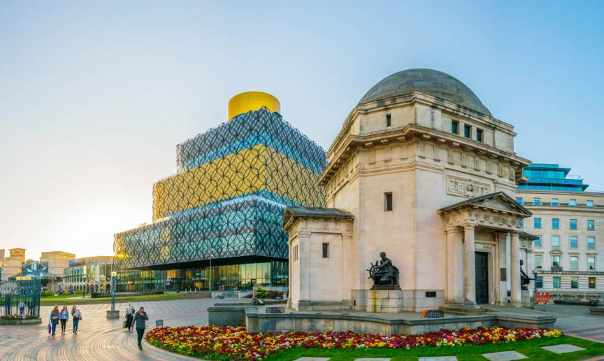 Stone built memorial hall with a small well kept and vibrant garden central to a large pedestrian walkway. In the background a four storey rectangular building with geometric pattern on the outside, alternating between a gold and blue glass colour scheme.