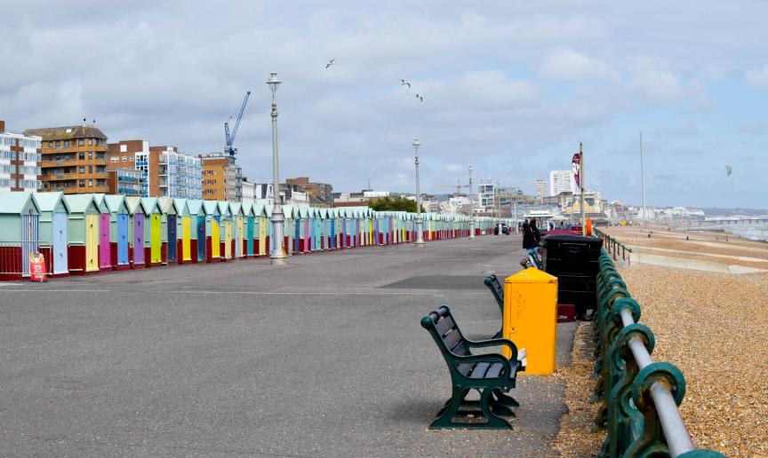 Concrete promenade walkway, to the left lined with a bold multi-coloured petite beach huts and to the right a shingled beach with multiple benches for a sea view.