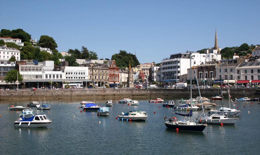 A busy harbour with numerous smaller boats decoratively displaying colourful buoys. A mix of properties cascade down from the hills towards the harbour.