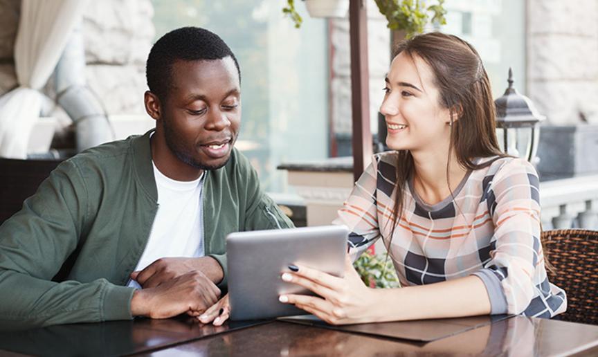 A male being shown something on a tablet style device by a woman, whilst sat next to each other at a table