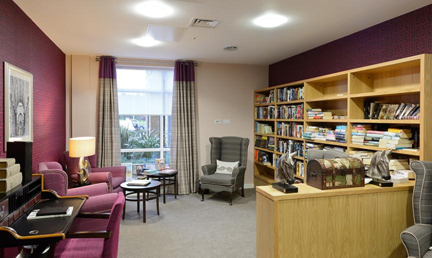 Four tall wooden oak bookcases fill the wall in the library, as well as four waist height shelving units to create a book corner. All of the shelving are adorned with books. The library area has scattered armchair seating. 