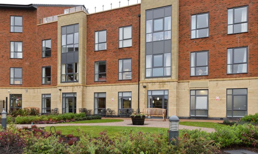 The four storey retirement community. The base of the property is built all in yellow brick, the further three storeys are built using a reddish brown brick with the protruding properties also built with the yellow bricks producing vertical stripes up the property. A outdoor rooftop balcony is seen on the right hand side of the property.