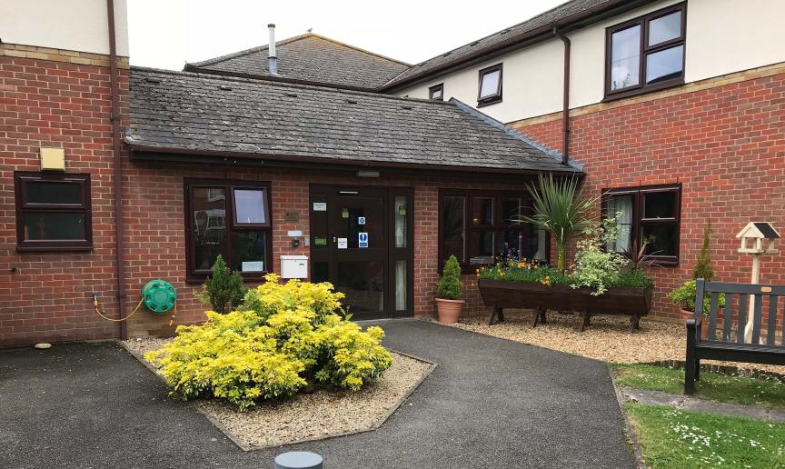 Multi elevational supported living property, built from red brick and a white rendered top elevation. A secure wooden leads into the property surrounded by mature shrubbery and large wooden planters