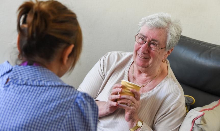 A resident sat in a chair holding a drink and smiling at a relative who has come to visit
