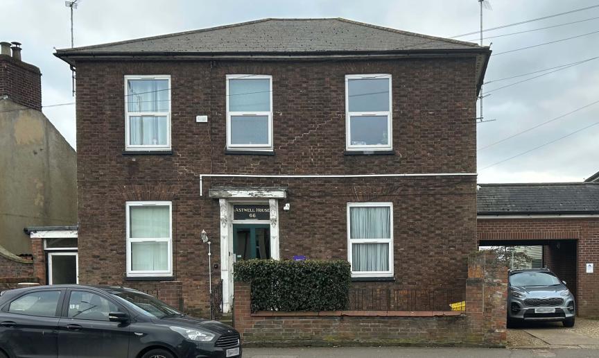 A detached, two storey brown brick built property, with a small brick built wall around the perimeter of the main building. Joined onto the building is a covered opening housing a parking space and access to the back of the property. 