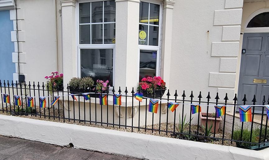 A close up image of the exterior of the building with a grey front door, bay window with sash windows and a black picket style fence out of the front with rainbow pride flags