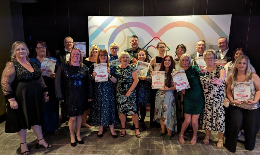 A group of 19 people standing together and smiling at the camera, wearing party outfits and suits, holding up certificates and awards.