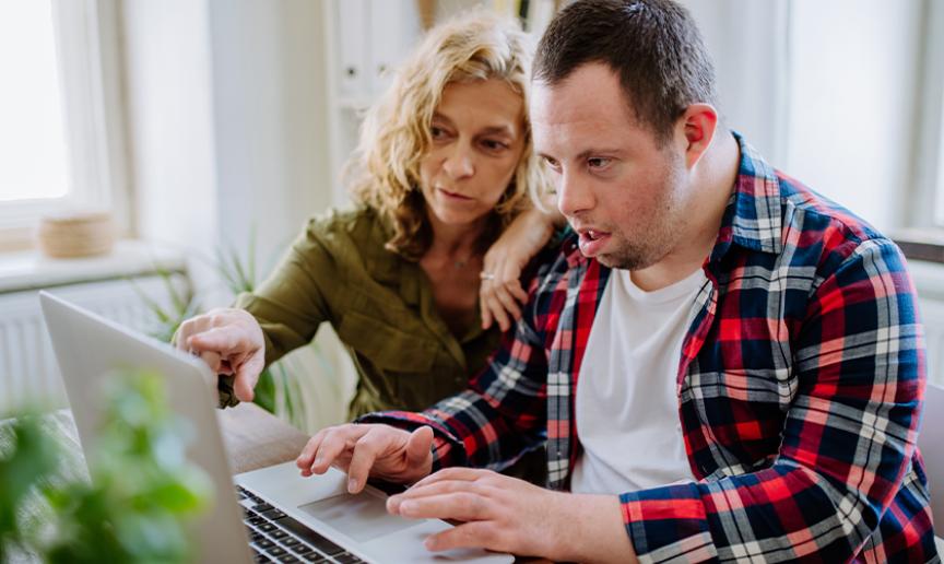 Stock photo of a young man with down syndrome wearing a red chequered shirt using a laptop on a wooden table with the help of an older woman wearing a green shirt