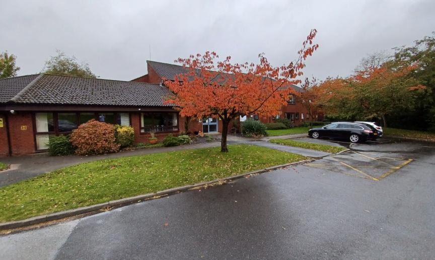 Exterior of red-brick building with a black tiled roof with grass, trees and parking spaces in front