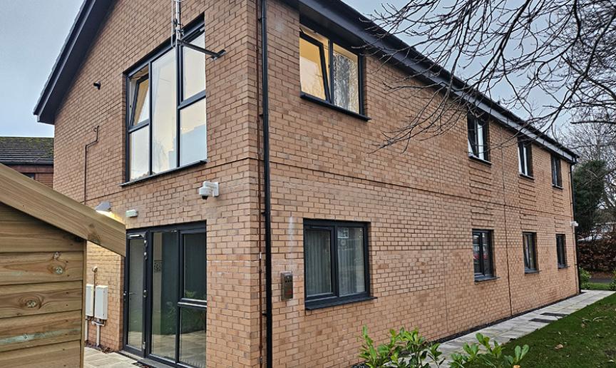 Exterior view of a two-storey supported housing building with light brown brick walls, large black-framed windows, and a paved pathway surrounded by greenery.
