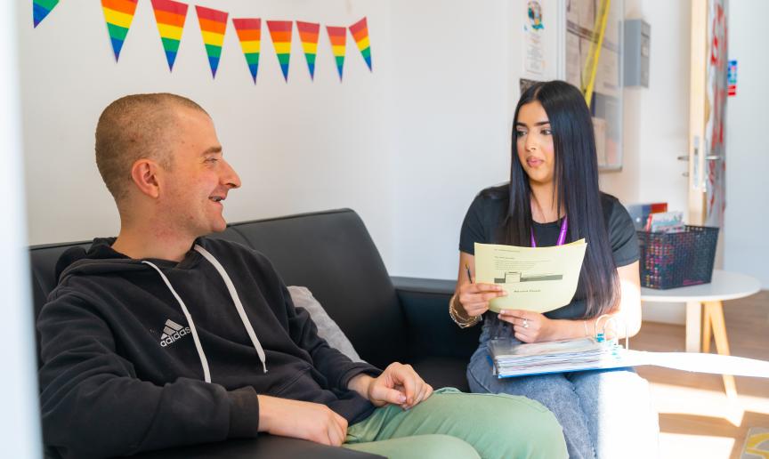 Two young people sat on a sofa one has a binder and is holding some papers