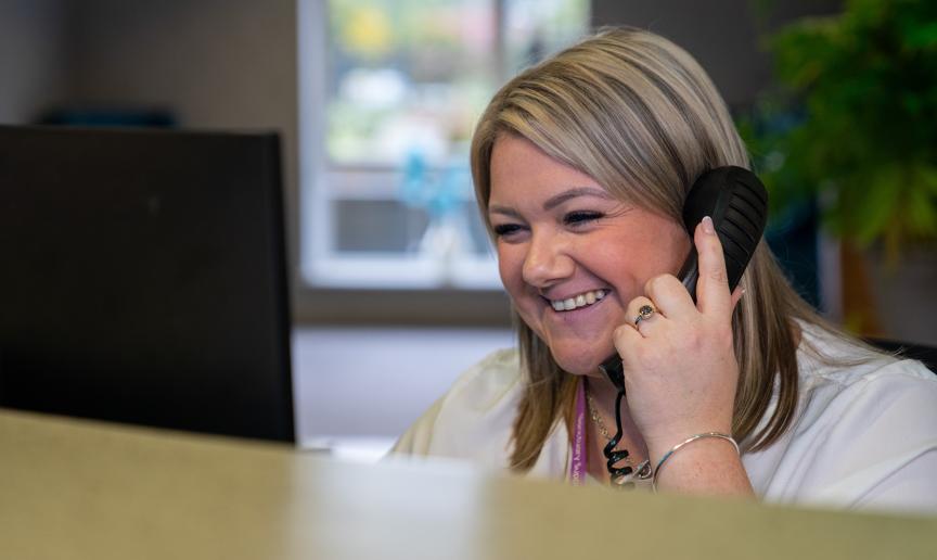 A person with smiling face is sitting at a desk, holding a phone to their ear. The person is in an office setting with a computer monitor in front of them and a window in the background. There is also a plant visible on the right side of the image.
