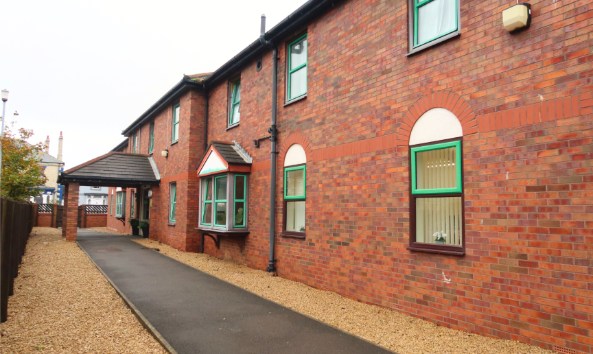 A brick building with green window frames and a sloped roof. The area is surrounded by gravel and a wooden fence, with a cloudy sky overhead.