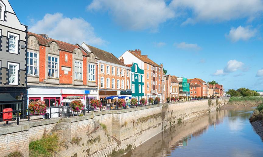 A row of colorful historic buildings along a riverside promenade on a bright sunny day, with reflections in the water