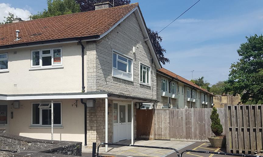 A two-story residential building with a light-colored exterior and a red-tiled roof is visible. In front of the building, there is a gated entrance leading to a parking area. The surrounding area features a stone wall and a wooden fence, with trees and greenery in the background. The sky is clear with a few clouds.