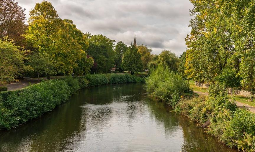 A calm river bordered by lush green trees and shrubs, with a church spire visible in the distance under a cloudy sky