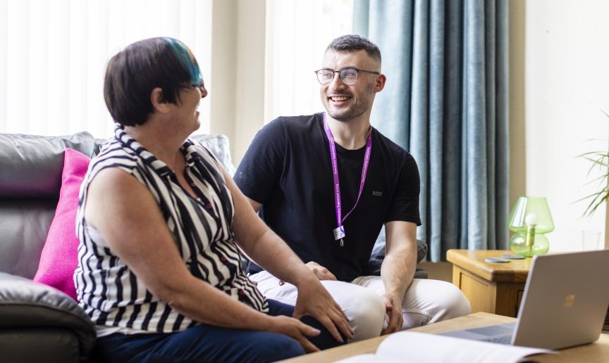 A resident and a Sanctuary Supported Living staff member are sitting on a soft couch together with a laptop on the coffee table