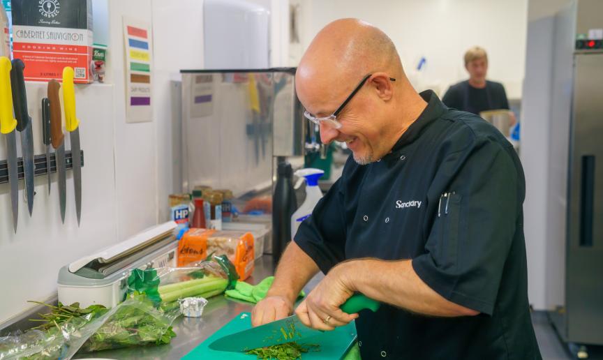 A chef with glasses is chopping herbs on a green cutting board in a modern kitchen. In the background, another person is visible, engaged in a different cooking activity.