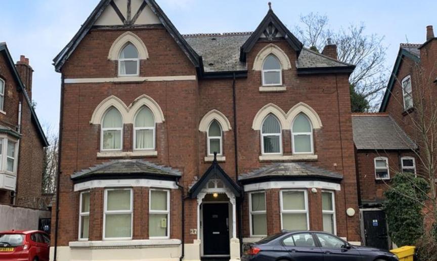 A large, three-story Victorian-style brick house with decorative gables and arched windows. The building features a symmetrical facade with a central entrance and two large bay windows on either side. The roof has a steep pitch, and there are trees in the background. A parked car is visible in front of the house, and the sky is partly cloudy.