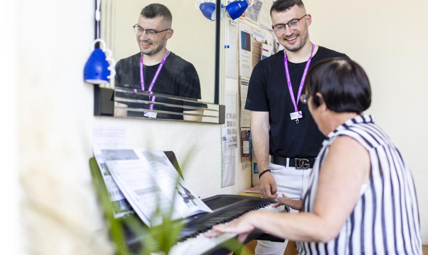 A staff member smiles while conversing with a resident while the resident plays piano in a brightly lit room