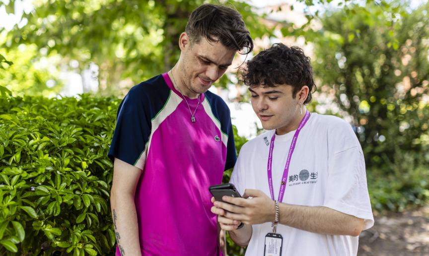 A Sanctuary Supported Living employee and a young male stood together looking at a phone outside in the sunshine