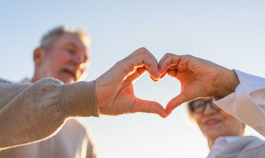 Two older people are making a heart shape with their hands, it is a bright and clear day
