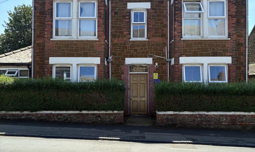 A 3 story dark brown brick building with white window panes. A hedge surrounds the front perimeter with a gap in the middle to allow entry