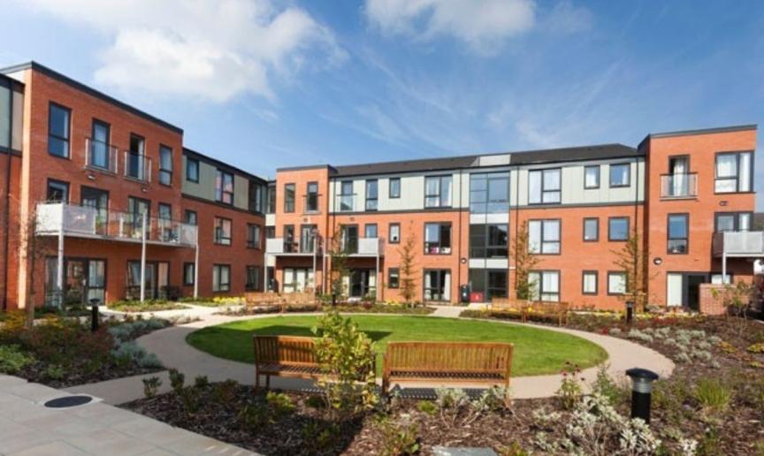 A modern, multi-story brick apartment building with large windows sits under a bright blue sky with scattered white clouds. A circular garden with a well-maintained green lawn and flowerbeds surrounds the building, featuring a wooden bench and pathway.