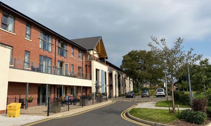 A street scene featuring a row of modern, multi-story brick apartments under a cloudy sky. A paved road curves gently ahead, with several parked cars and a yellow traffic cone visible. Trees and landscaping flank the road, and a small seating area is present at the front of the buildings.