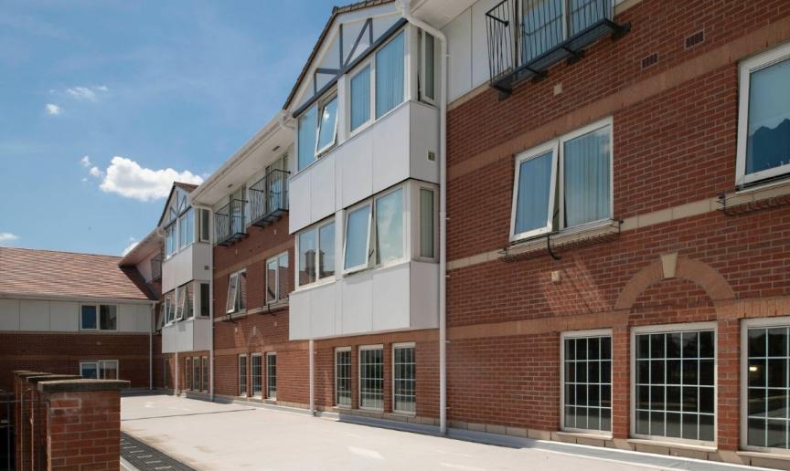 Exterior view of a multi-story brick building under a bright blue sky. The building features several windows with balconies on the upper floors, and a paved courtyard extends to the left. A decorative stone pillar is visible in the foreground.