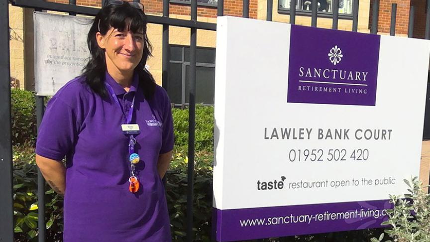 Wellbeing and Inclusion Assistant stands in front of the sign for Lawley Bank Court, in a purple Sanctuary uniform top