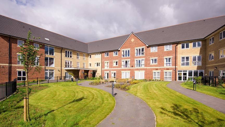 The newly built, yellow, and red bricked, three storey retirement living property set behind winding pathways which lead to a circular outdoor seating area as well the property and lovely grassed areas.