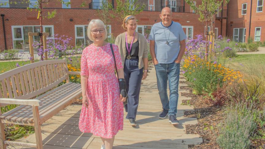 Three people walk in a sunny garden, on the left a happy older woman with glasses and short white hair wearing a pink floral dress. In the centre, a woman working for Sanctuary with brown hair, wearing a casual outfit. On the right, a cheerful man wearing a casual grey shirt and jeans. The garden is filled with colourful flowers and greenery, creating a vibrant atmosphere.