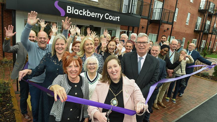 A group of people gathered outside a modern brick building with balconies, standing in front of a sign that reads “Blackberry Court.” They are participating in a ribbon-cutting ceremony, holding a long purple ribbon stretched across the entrance. Many individuals have their hands raised in celebration, and the setting appears to be the opening of a new facility or building.