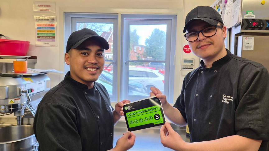 Two chefs in black uniforms and caps are standing in a kitchen. They are smiling and holding up a green food hygiene rating certificate that displays a score of 5. The kitchen is equipped with various cooking appliances and utensils, and there is a window in the background showing a view of parked cars outside.