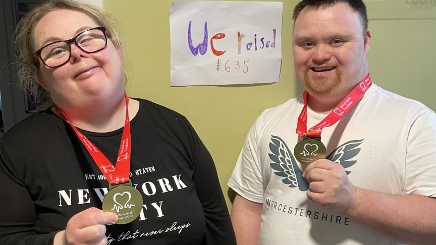 A woman and a man stand side by side, both smiling and holding medals. They are wearing red lanyards around their necks. Behind them is a sign that reads "We raised 1635," indicating a fundraising achievement.