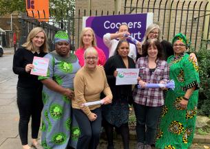 A group of nine women standing together in the street by a large sign that reads 'Coopers Court', smiling at the camera and holding signs that read 'FareShare'.