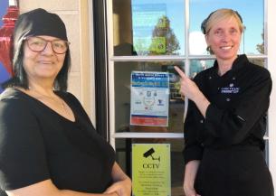 Two women smiling at the camera, pointing to a sign on a door that shows a rating of 5 from the Food Standards Agency.