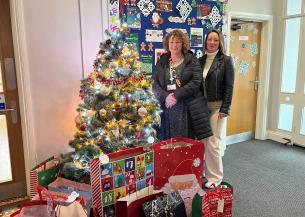 Two women stood by a Christmas tree and smiling at the camera, surrounded by gift bags containing presents.