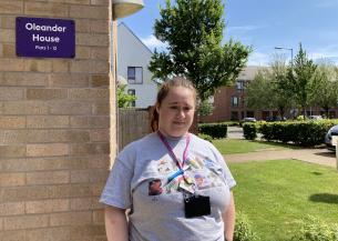 A woman who appears to have Down syndrome, wearing a grey t-shirt and a purple work lanyard, standing in front of a building with greenery in the background