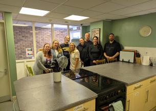  A group of eight white men and women in their 20s, 30s and 40s, standing together and smiling at the camera. They're stood in a kitchen with freshly-painted green walls. In front of them is a kitchen island with two cookers and a pot of utensils. They're all smiling at the camera.