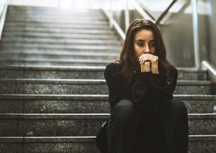A woman in her 20s wearing dark clothes and with long dark hair, sat on the dark stairs like those which lead down to an underground station or walkway. She looks worried and has her hands clenched in front of her face.