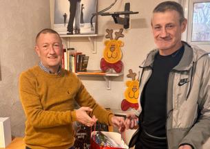 Two men standing together, the first wearing a mustard coloured jumper and dark wash jeans, and the second, taller man in a Nike tracksuit top. They are holding Christmas gift bags and smiling.