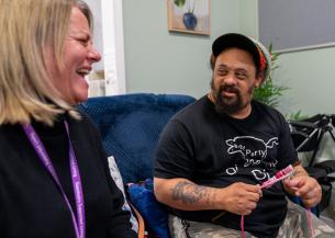 A white woman with blonde hair wearing a purple Sanctuary Supported Living lanyard laughs next to a man with darker skin, wearing a rastacap, sideburns, and a beard, using crochet needles with pink and white wool