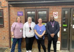 Three white women and one black man standing in a row outside by the front door of a residential care home. They are all wearing purple lanyards and are smiling at the camera. There is a green sticker in the window behind them, showing a rating of five from the Food Standards Agency.