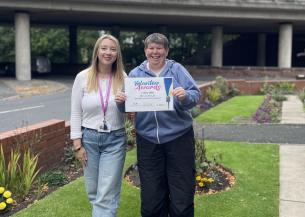 two women standing together, smiling and looking at the camera. A blonde woman on the left is wearing a white top and a purple lanyard, the woman on the right is holding up a certificate that reads volunteer awards.