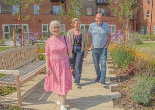 Three people walk in a sunny garden, on the left a happy older woman with glasses and short white hair wearing a pink floral dress. In the centre, a woman working for Sanctuary with brown hair, wearing a casual outfit. On the right, a cheerful man wearing a casual grey shirt and jeans. The garden is filled with colourful flowers and greenery, creating a vibrant atmosphere.